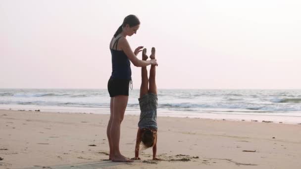 Sportieve jonge moeder met dochtertje gymnastische doen op het strand. — Stockvideo