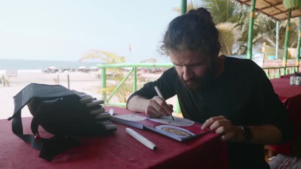 El hombre se sienta en la cafetería y dibuja la imagen de la comida india en el cuaderno de bocetos con marcadores de arte — Vídeos de Stock