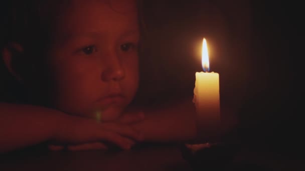 Портрет маленькой милой девочки, которая сидит в темноте и смотрит на горящую свечу — стоковое видео