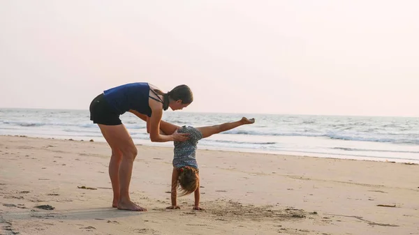 Joven madre deportiva con hija pequeña haciendo gimnasia en la playa . — Foto de Stock