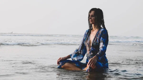 Jovem mulher molhada sentada na água na praia de areia e fazendo práticas de meditação — Fotografia de Stock
