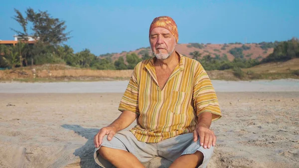 Senior man sitting and meditating on sandy beach with crossed feet — Stock Photo, Image