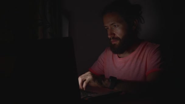 Jonge, bebaarde man aan het werk op laptop in duisternis — Stockvideo