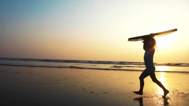 Menina está correndo ao longo da praia de areia do oceano com prancha de surf ao pôr do sol . — Vídeo de Stock