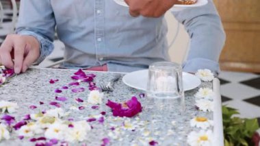 Waiter brings breakfast to a couple sitting at the table covered with flowers.