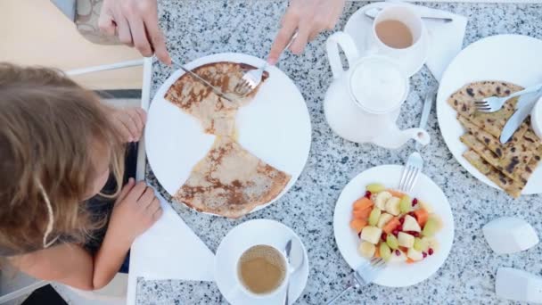 Mała dziewczynka jest jedzenie naleśniki na śniadanie. Moms ręce wyciąć je na kawałki — Wideo stockowe