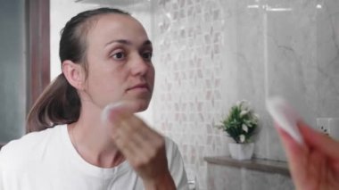 Genç bir kadın banyoda temizlik losyonuyla yüzünü pamukla siliyor..