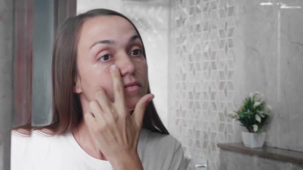 Jovem mulher está aplicando creme no rosto sob a área dos olhos no banheiro . — Vídeo de Stock