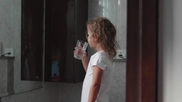 Little curly child girl rinses her mouth with water in bathroom, side view.