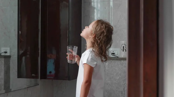 Küçük kıvırcık çocuk kız banyoda su ile ağzını durular, yan görünüm.