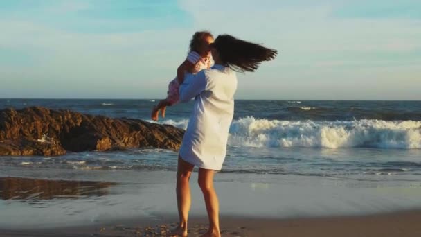 Mãe com rindo pequena filha em seus braços girando na praia do oceano de areia — Vídeo de Stock