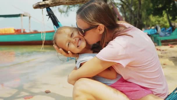 Mamá está abrazando y besando a su pequeña hija sentada en una hamaca en la playa del océano — Vídeos de Stock