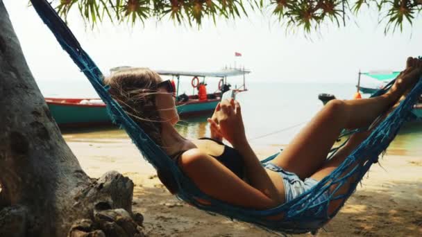 Woman is resting in hammock browsing pages in smartphone in ocean sand beach. — Stock Video