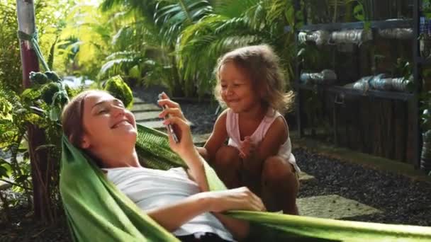 Mãe na rede com telefone está brincando com sua filhinha sentada perto dela . — Vídeo de Stock