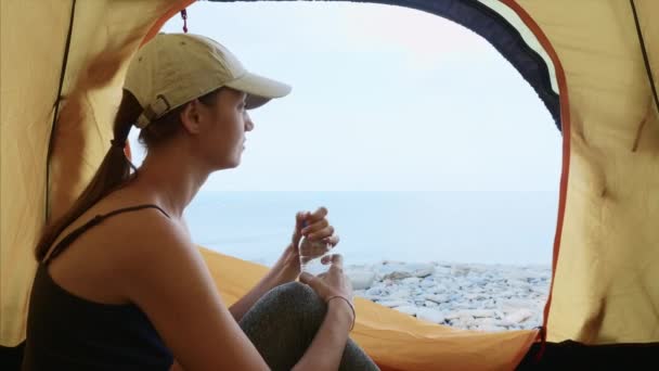 Woman is sitting in camping tent, drinking water from bottle and looking at sea. — Stock Video