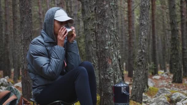 Wanderin in Jacke trinkt heißen Tee im Waldcamping auf Stuhl sitzend. — Stockvideo