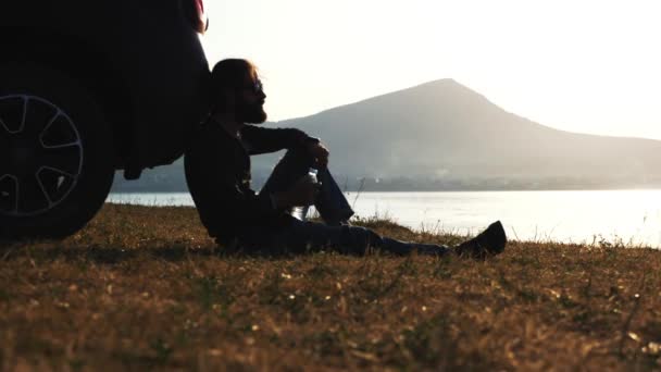 Young bearded man is sitting on the ground, leaned on his car and drinking water — Stock Video