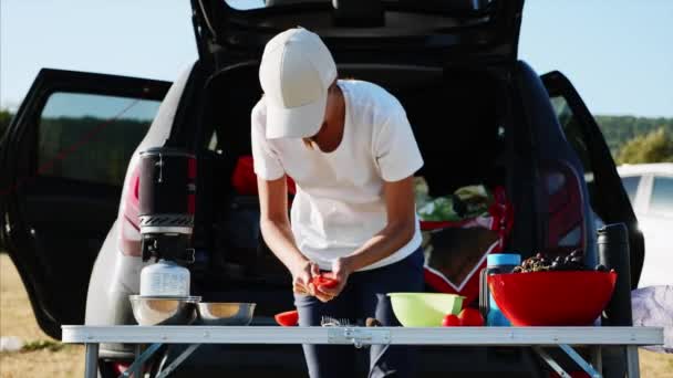 Jeune femme tranchant des tomates sur la table près d'une tente touristique au camping . — Video