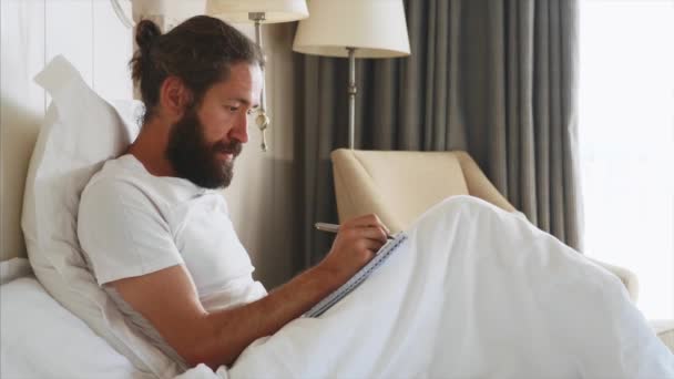 Pemuda berjanggut menulis catatan untuk buku harian setelah terbangun di kamar tidur di rumah — Stok Video