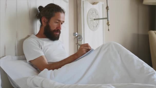 Νέος άντρας με γένια γράφοντας σημειώσεις στο ημερολόγιο μετά την ξύπνια στο υπνοδωμάτιο στο σπίτι — Αρχείο Βίντεο