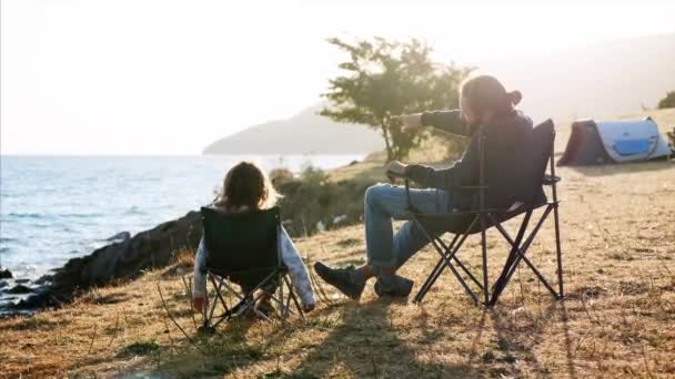 Father with little daughter sits on chairs at camping on lake shore, rear view — Stock Video