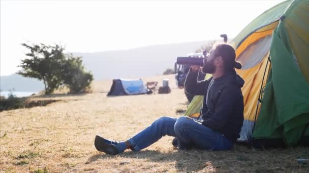 Young man sits on near the tent at campsite and drinking tea from thermos bottle — Stock Video