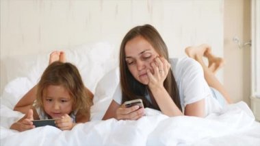 Küçük sevimli kızı ile genç anne beyaz bir yatakta yatan gadget'lar kullanıyor.