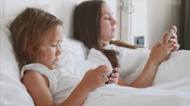 Küçük sevimli kızı ile genç anne beyaz bir yatakta yatan gadget'lar kullanıyor.