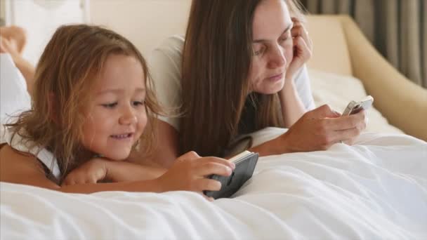 Joven madre con pequeña hija linda están utilizando gadgets acostado en una cama blanca . — Vídeo de stock