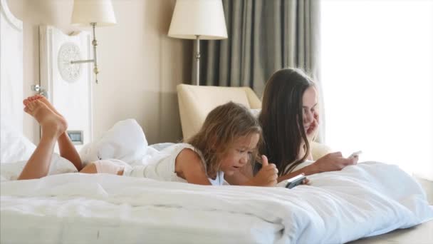 小さなかわいい娘を持つ若い母親は、白いベッドに横たわってガジェットを使用しています. — ストック動画