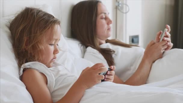 Jovem mãe com pouco bonito filha estão usando gadgets deitado em uma cama branca . — Vídeo de Stock