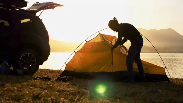 Silhouette of young bearded man sets up a tent at campsite on the lake shore. — Stock Photo, Image