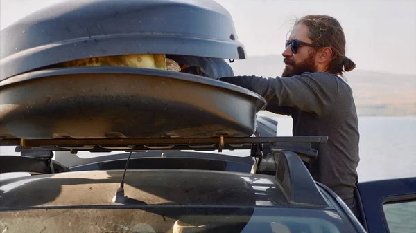 Genç sakallı adam araba çatı kutusundan bir kamp ekipmanları dışarı alıyor