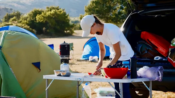 Mujer joven rebanando tomate en la mesa cerca de una tienda de campaña turística en el camping . — Foto de Stock