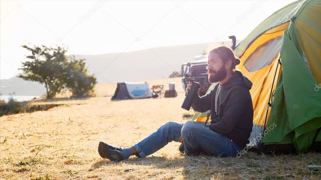 Young man sits on near the tent at campsite and drinking tea from thermos bottle