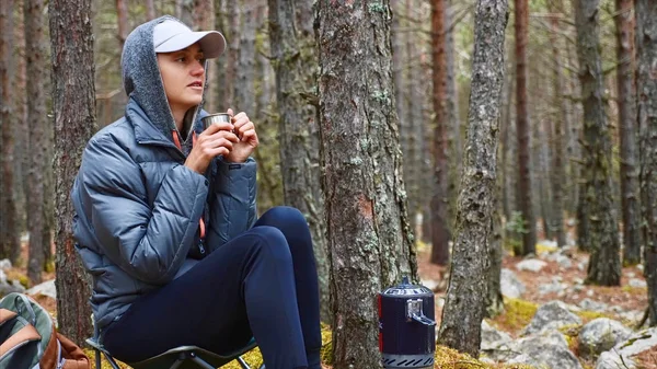 Mujer excursionista en chaqueta bebe té caliente en bosque camping sentado en silla . — Foto de Stock