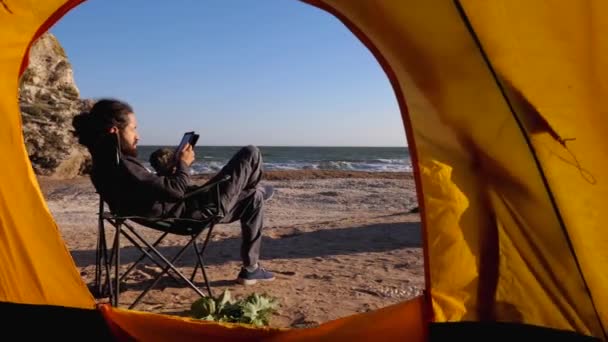 Widok z namiotu kempingowego do brodatego człowieka czyta książkę elektroniczną na plaży — Wideo stockowe