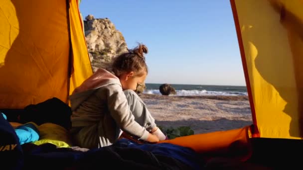 Anak perempuan menempatkan sepatunya sementara duduk di tenda berkemah di pantai yang indah — Stok Video