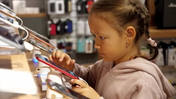 小さな子供の女の子は、電子店でカウンターの上に新しいスマートフォンを使用して. — ストック動画