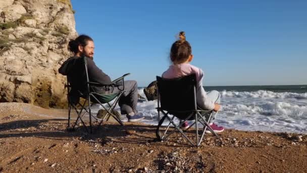 Батько з маленькою донькою сидить на стільцях на пляжі, вид ззаду . — стокове відео