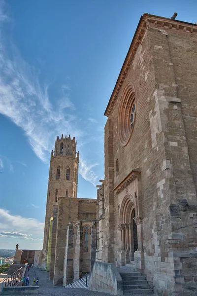 Katalonya 'nın eski katedralinin kulesi, yaz güneşiyle aydınlanan taş duvarlardan görkemli bir şekilde yükselir. Yoğun mavi gökyüzü, ortaçağ Gotik mimarisi, tarihi aktarma ve anıtsallığı ile zıtlığı güçlendiriyor.