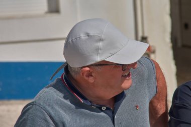 Orta yaşlı bir adam gülümsüyor ve rahat görünüyor, açık gri bir şapka ve koyu güneş gözlüğü takıyor, yanında da mavi renkli polo gömleği var. İçerik ifadesi açık havada güneşli bir günün tadını çıkardığını gösteriyor. T