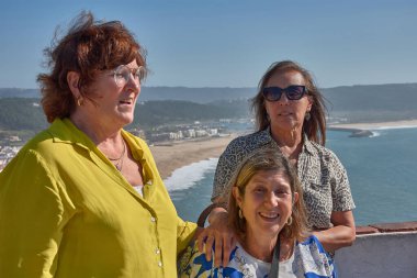 Portekiz, Nazare 'de üç orta yaşlı kadın poz veriyor. Resimli sahil kasabası, sahil ve arka planda açık mavi bir gökyüzünün altında geniş Atlantik Okyanusu var..