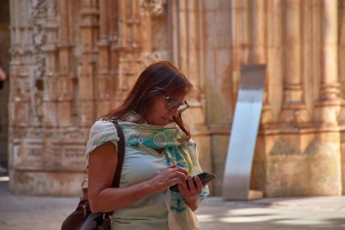 Gözlük takan olgun bir kadın ve desenli bir eşarp cep telefonunu kontrol ederken odaklanmış görünüyor. Portekiz 'deki Batalha Manastırı' nın içini ya da çevresini hatırlatan bir çantasıyla omzunun üzerinde duruyor.