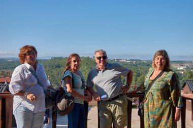 Portekiz 'in Obidos kentindeki ahşap yürüyüş yollarında poz veren ve sohbet eden bir grup emekli erkek ve kadın görülüyor. Bazıları kameraya gülümserken, diğerleri birbirleriyle etkileşime girer.