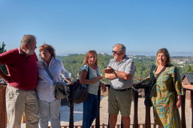 Portekiz 'in Obidos kentindeki ahşap yürüyüş yollarında poz veren ve sohbet eden bir grup emekli erkek ve kadın görülüyor. Bazıları kameraya gülümserken, diğerleri birbirleriyle etkileşime girer.