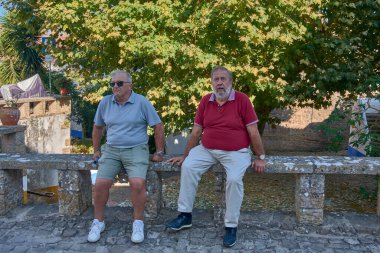 İki emekli arkadaş, orta yaşlı adamlar, bir taşta ya da ahşap bir bankta oturuyorlar, rahatlar ve açık havada canlı canlı sohbet ediyorlar.. 
