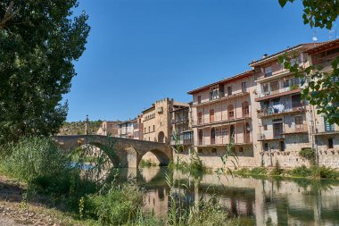 Valderrobres 'in tarihi taş köprüsü, Teruel, Matarraa Nehri boyunca uzanan, arka planda ortaçağ kasabasının heybetli giriş kulesi. Güneş ışığı sahneyi aydınlatıyor, inşaatın sağlamlığını vurguluyor.