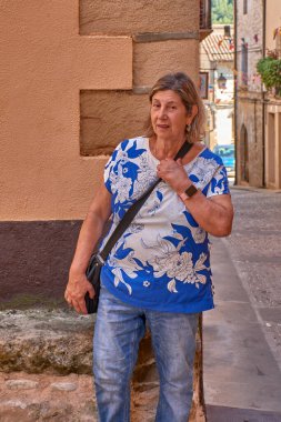 Beyaz ve mavi baskılı tişört ve kot pantolon giyen bir kadın kaldırım taşlı bir köy caddesinde doğal ve rahat bir ifadeyle poz veriyor. Güneş ışığı yüzünü ve vücudunu aydınlatırken, koyu tonlu binaların geleneksel mimarisi... 