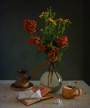 Masanın üzerinde bir fincan aromatik kahve ve kitap ayracı yerine kağıt tekne olan açık bir kitap var. Bir buket kırmızı böğürtlen ve vazoda çayır çiçekleriyle dolu bir buket Rowan dalı. Durgun hayat. Eski.